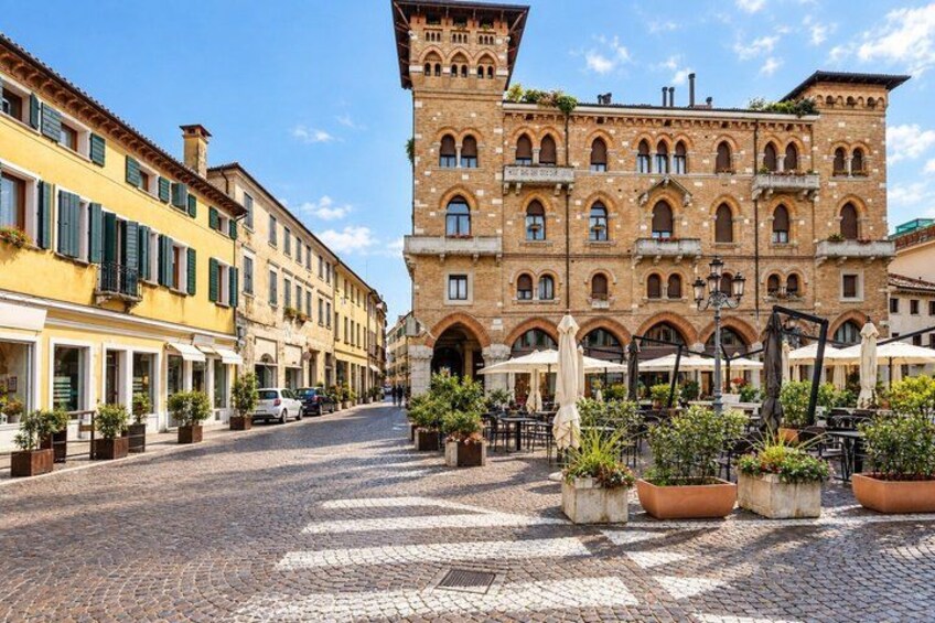 A cobblestone street with buildings, outdoor seating, and potted plants. 3