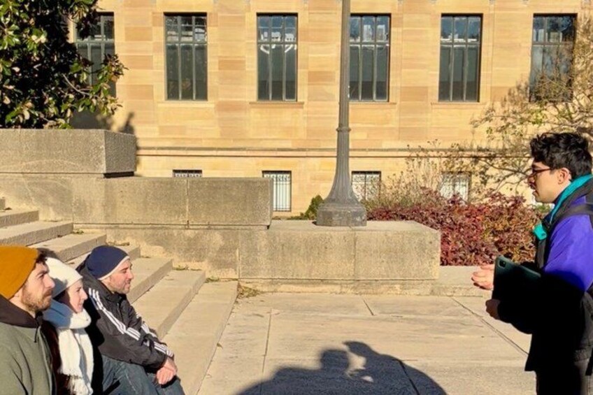 A person standing and gesturing in front of a building with steps and a tree. 2