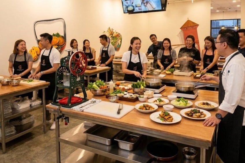 A group of people in a kitchen setting, some wearing aprons, preparing food on tables with various dishes and cooking utensils. 1