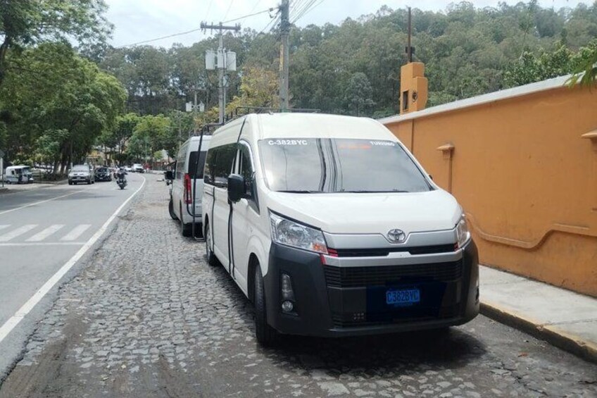 A white van parked on a cobblestone street with a yellow wall on the right. 4