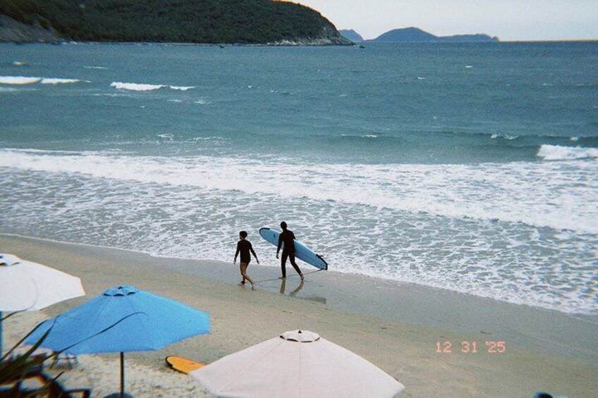 A beach with two people walking with surfboards, umbrellas, and a date stamp. 5