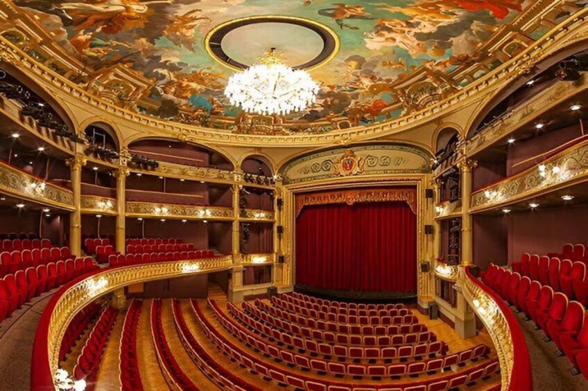 An ornate theater interior with red seats, a grand chandelier, and a detailed ceiling fresco. 2