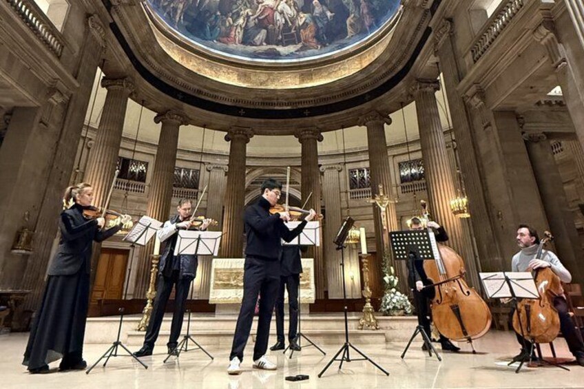 A classical music orchestra performing in a grand hall with columns and a large frescoed ceiling. 2