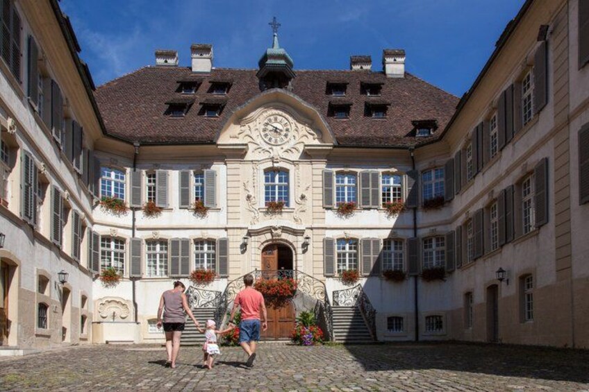 A cobblestone courtyard with a historic building featuring a clock tower, flower boxes, and a staircase leading to the entrance. 5
