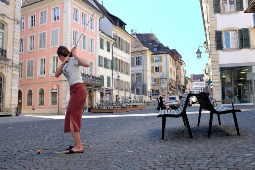 A person playing croquet on a cobblestone street, with historic buildings and benches in the background. 1