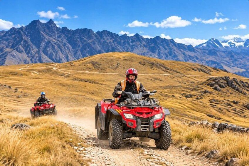 Two riders on red ATVs on a dirt trail with mountains in the background. 1