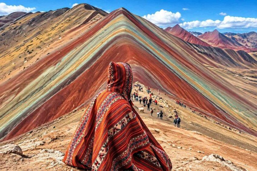 A person in traditional attire sitting on a rocky outcrop, overlooking a colorful mountain range with a group of people in the background. 2