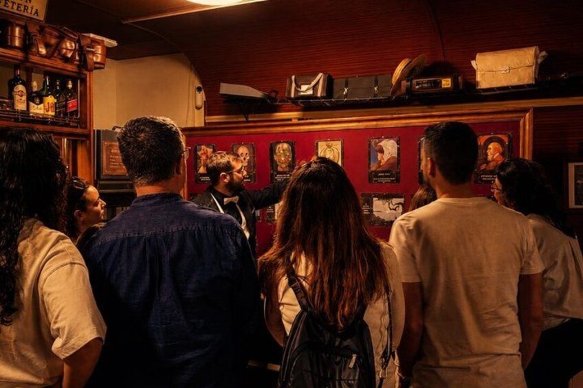 A bar with patrons, wooden shelves with bottles, and framed pictures on the wall. 1