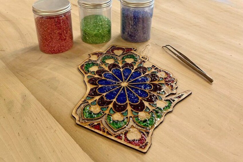 A wooden decorative piece with a blue and green mosaic pattern, three jars of colored glitter, and a pair of tweezers on a wooden surface. 5
