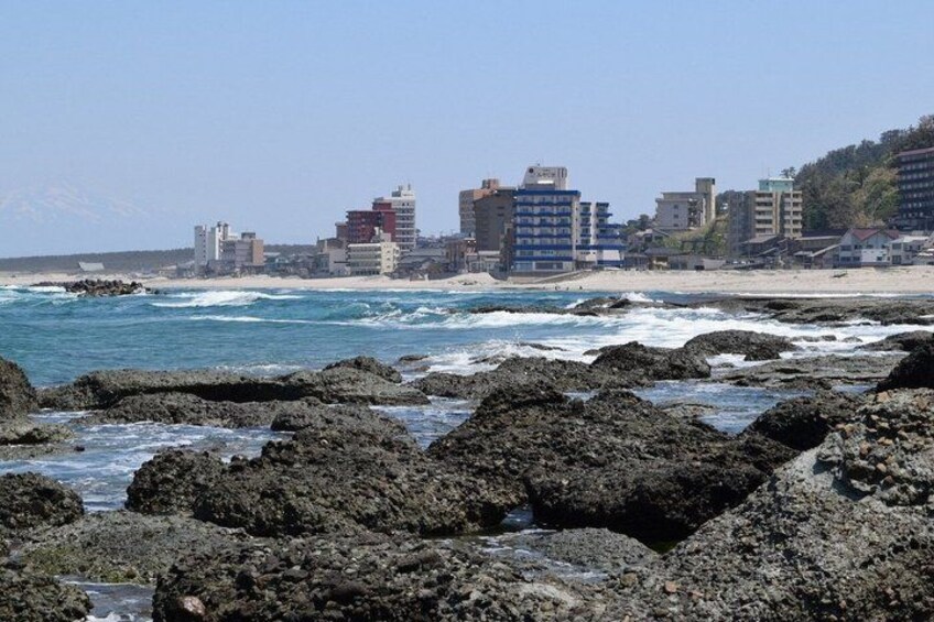A rocky shoreline with waves, a sandy beach, and buildings in the background. 1