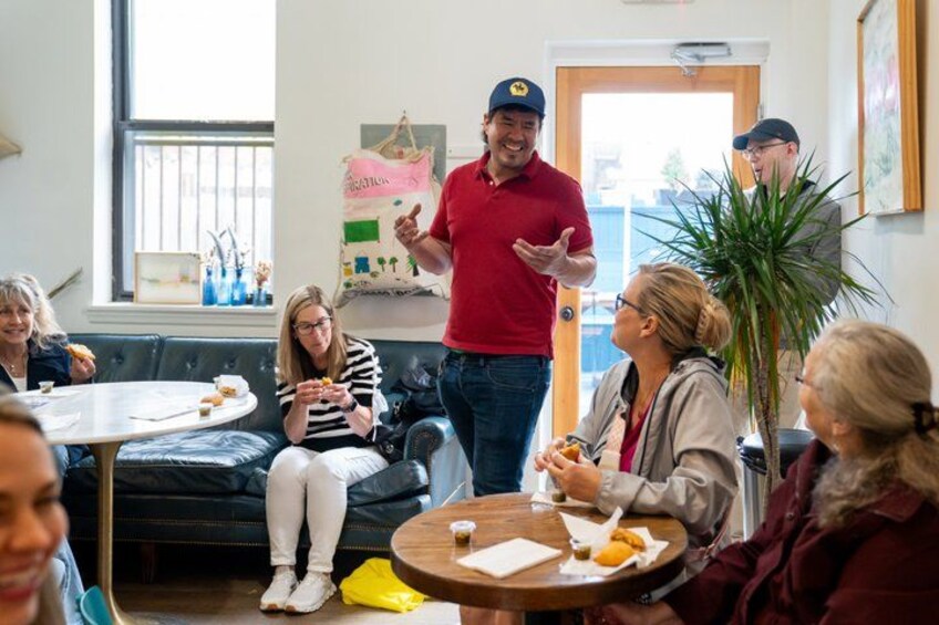 A group of people sitting at a table, eating and conversing in a room with a plant and a poster on the wall. 1
