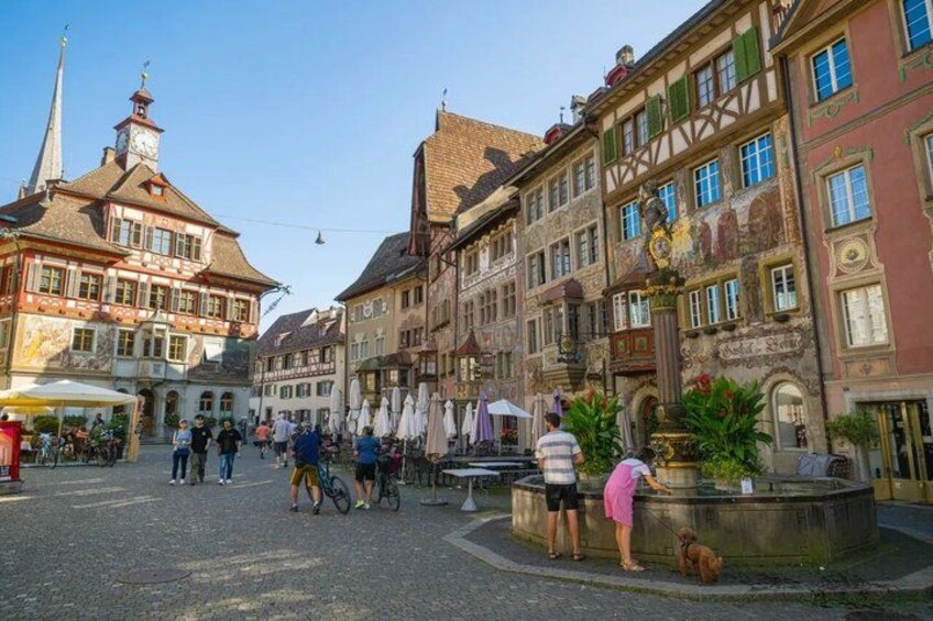 A cobblestone street with historic buildings, a fountain, and people walking and cycling. 2
