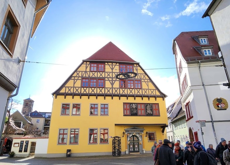 A yellow half-timbered building with a red roof, a black circular sign, and a crowd of people in front. 2