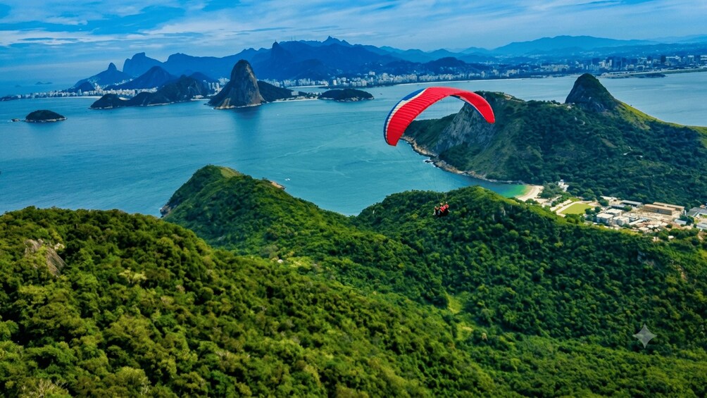 Un parapentista vuela sobre un paisaje costero con abundante vegetación y montañas al fondo. 1