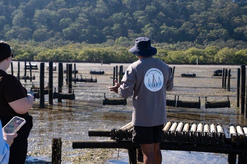 A person in a hat and jacket standing by a waterway with a boat, another person holding a device, and a forested area in the background. 5