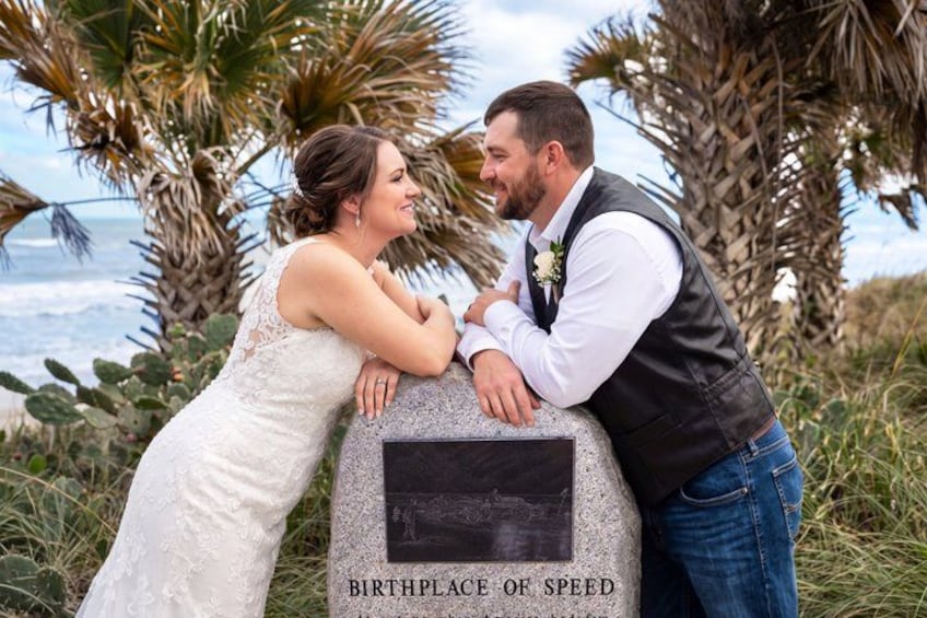 A couple leaning on a stone monument with the inscription 'BIRTHPLACE OF SPEED' in a coastal setting with palm trees and ocean in the background. 3