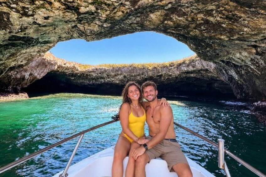 A boat with two people, a rocky cave entrance, and clear blue water. 1