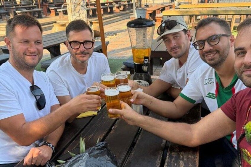 Four people toasting with beer glasses at an outdoor table with a blender. 4