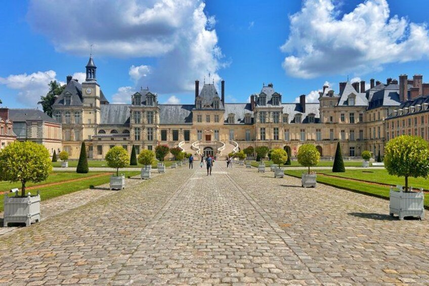 A historic building with a cobblestone courtyard, symmetrical landscaping, and a clear sky. 1