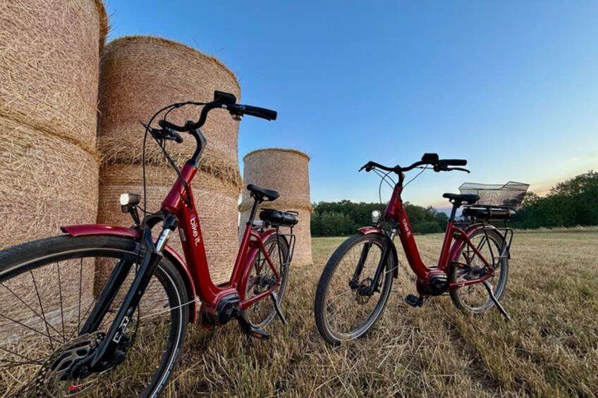 Two red bicycles parked in a field with hay bales in the background. 1