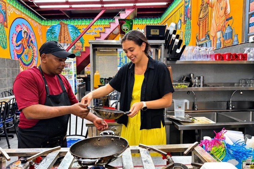 A person cooking in a kitchen with a wok on a stove, another person standing nearby, and colorful murals on the walls. 4