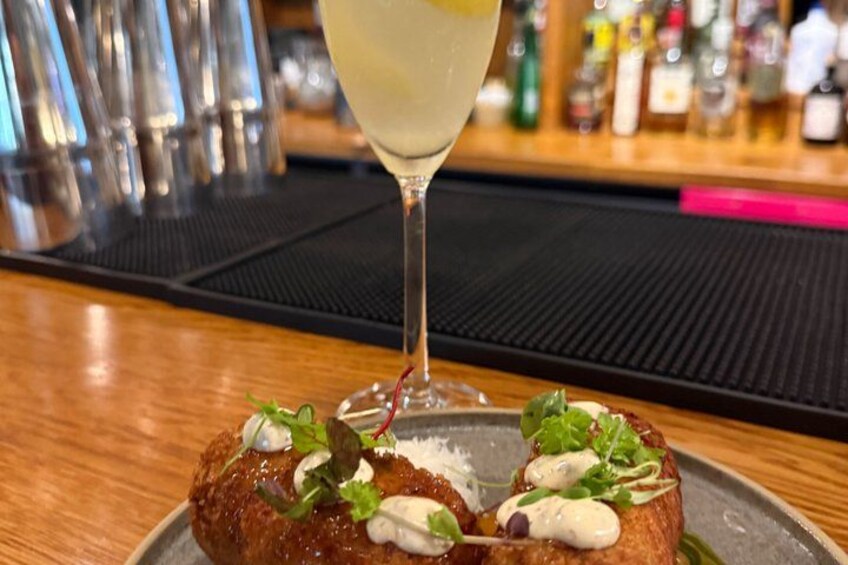A cocktail glass with a lemon wedge, a plate with a food item garnished with greens and a creamy sauce, and a bar setting with bottles in the background. 5