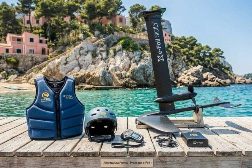 A blue life jacket, a helmet, a paddleboard, and a portable e-foil surfboard on a wooden dock by the sea. 3