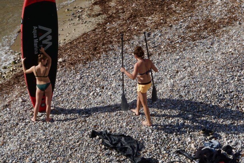 A pebbled beach with two people, one holding a surfboard and the other holding a paddle. 4