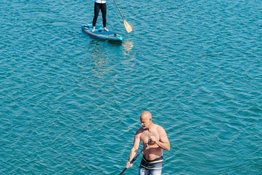 A person paddleboarding on a calm blue sea, another person standing on a paddleboard nearby, and a third person in a boat. 1