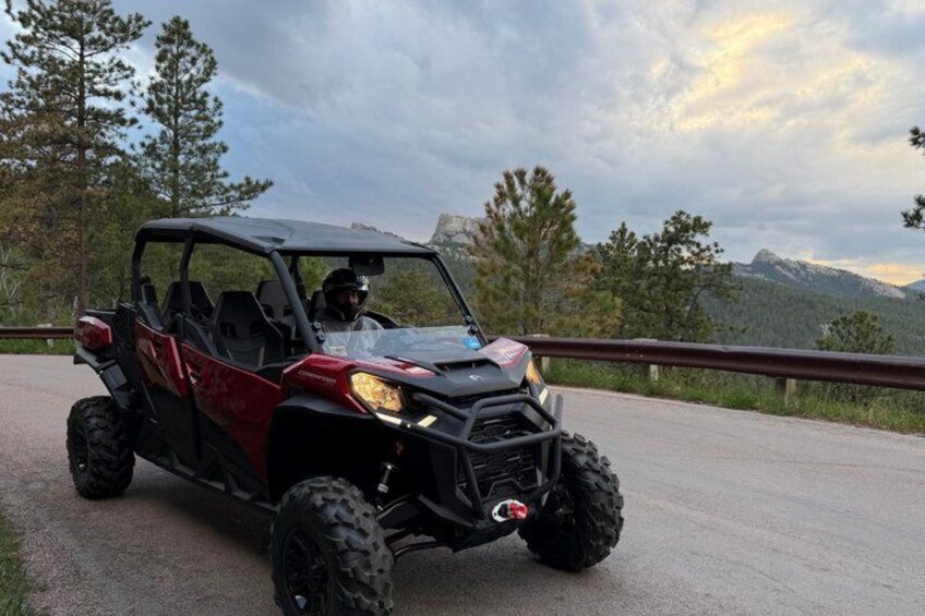 A red off-road vehicle on a paved road with a guardrail, surrounded by trees and mountains in the background. 3