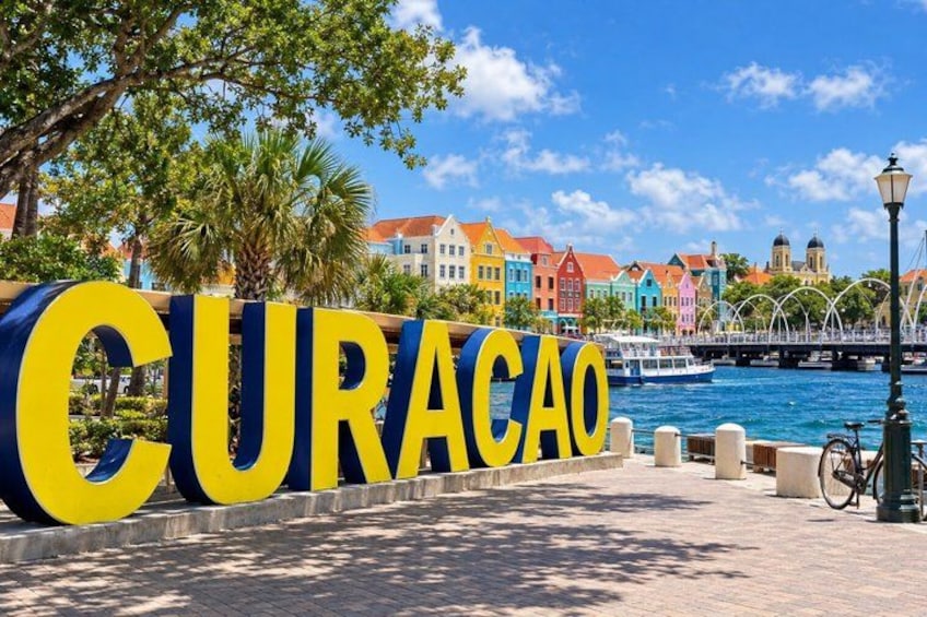 A large sign spelling 'CURACAO' in yellow and blue letters, with a waterfront and colorful buildings in the background. 5