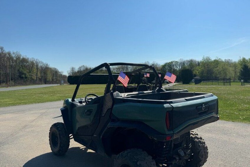 A green utility vehicle with American flags on the back, parked on a paved road with grass and trees in the background. 5