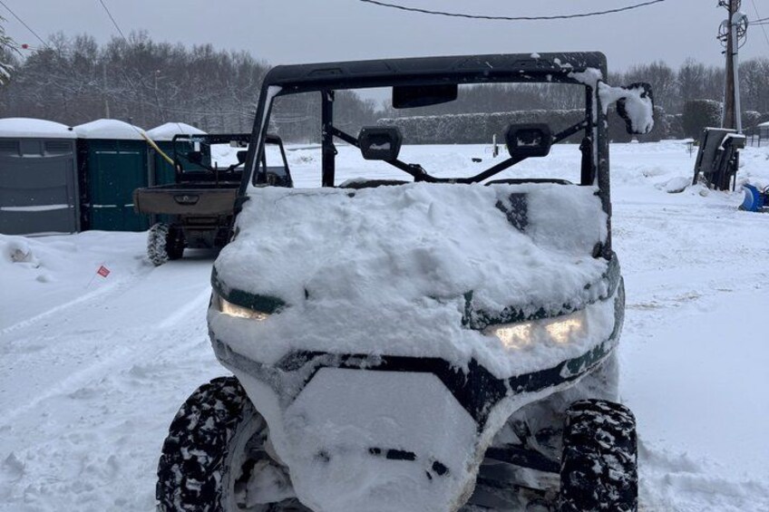 A snow-covered vehicle with a cabin, a snow-covered ground, and a background with trees and a building. 2