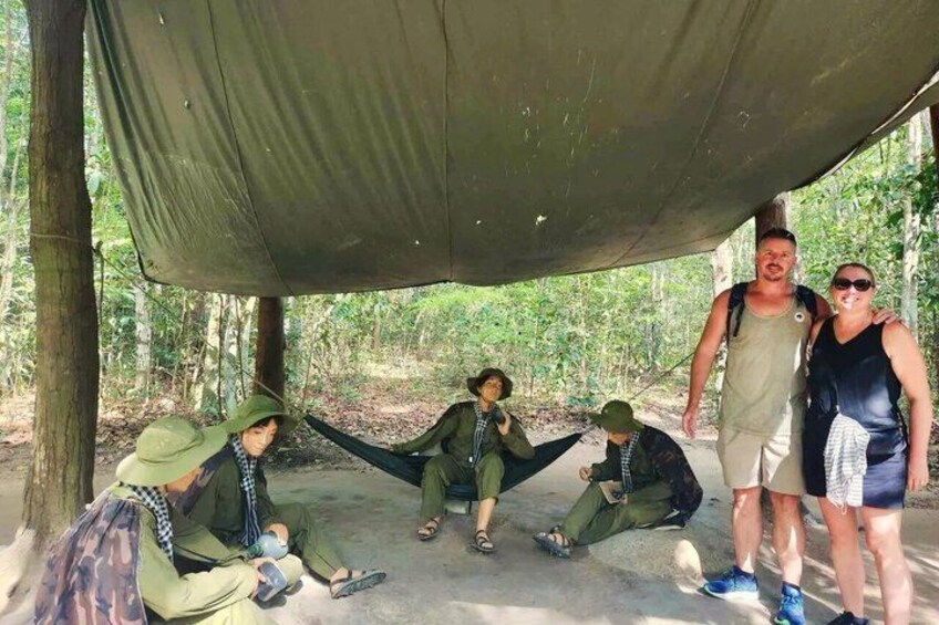 A group of people in a forested area under a canopy, some wearing hats and carrying backpacks. 2