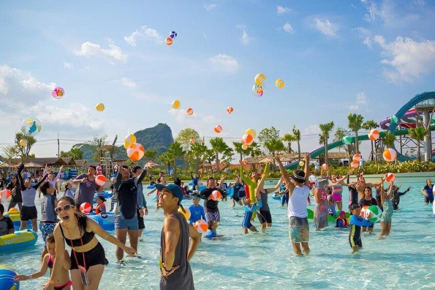 A crowded water park with people playing in the water, a water slide, and palm trees in the background. 3