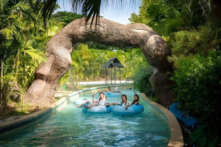 A pool with a large rock arch, people on inflatable tubes, and tropical vegetation. 1