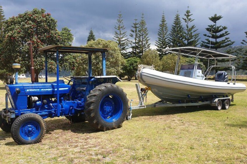 A blue tractor towing a boat on a trailer in a grassy field with trees in the background. 4