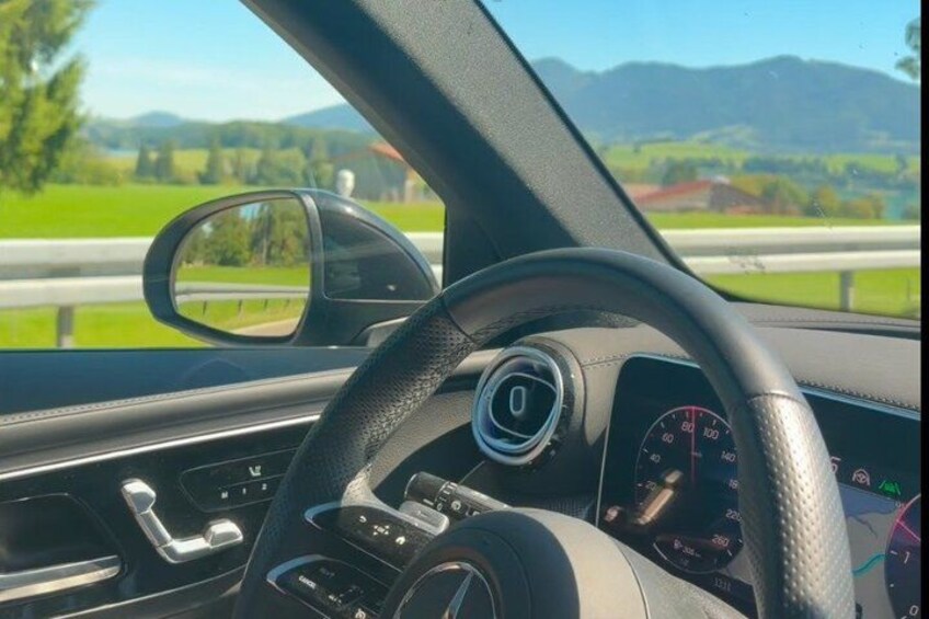 A car's interior with a steering wheel, dashboard, and a side mirror reflecting a road with greenery and mountains in the background. 5