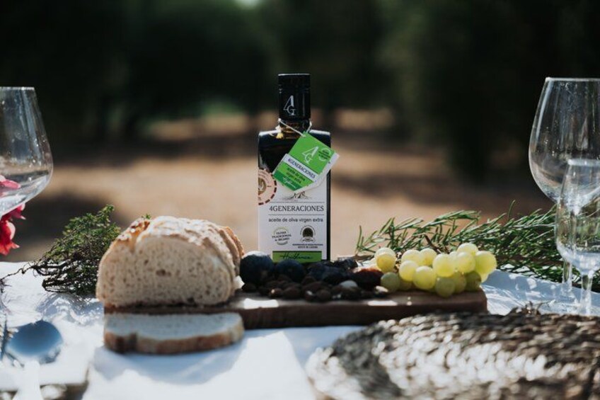 A cheese board with a bottle of 4 GENERACIONES olive oil, grapes, and bread on a table outdoors. 4