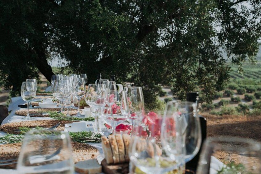 An outdoor dining table set with wine glasses, plates, and bread, surrounded by greenery and trees. 5