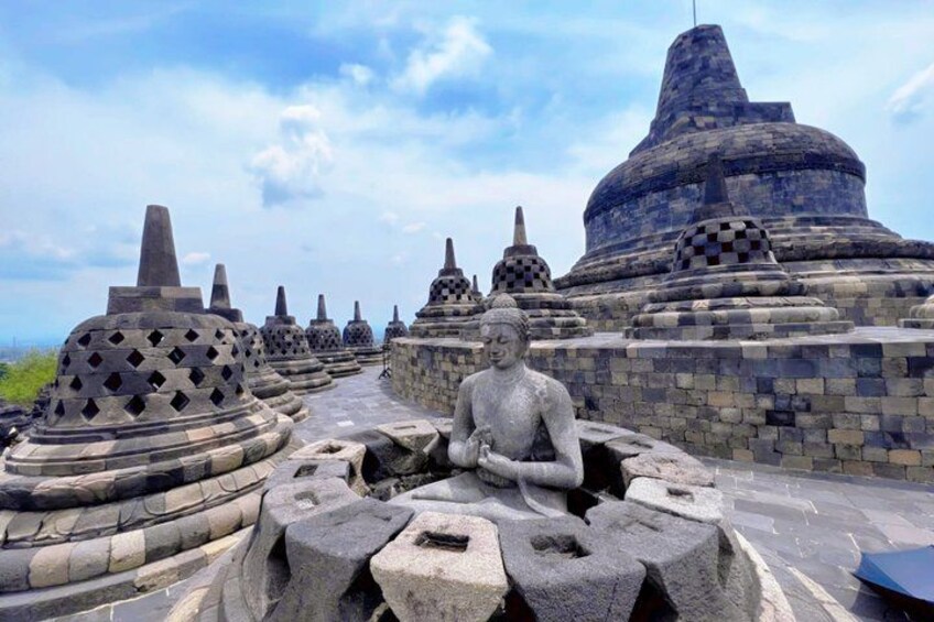 A stone Buddha statue in a meditative pose, surrounded by stone stupas and a large temple structure. 1