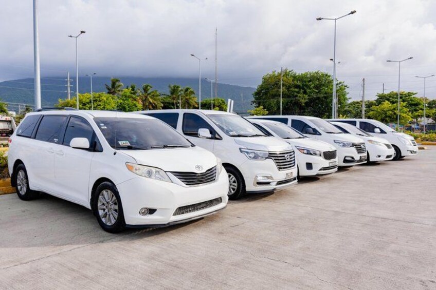 A row of white minivans parked in a lot with a cloudy sky and mountains in the background. 1