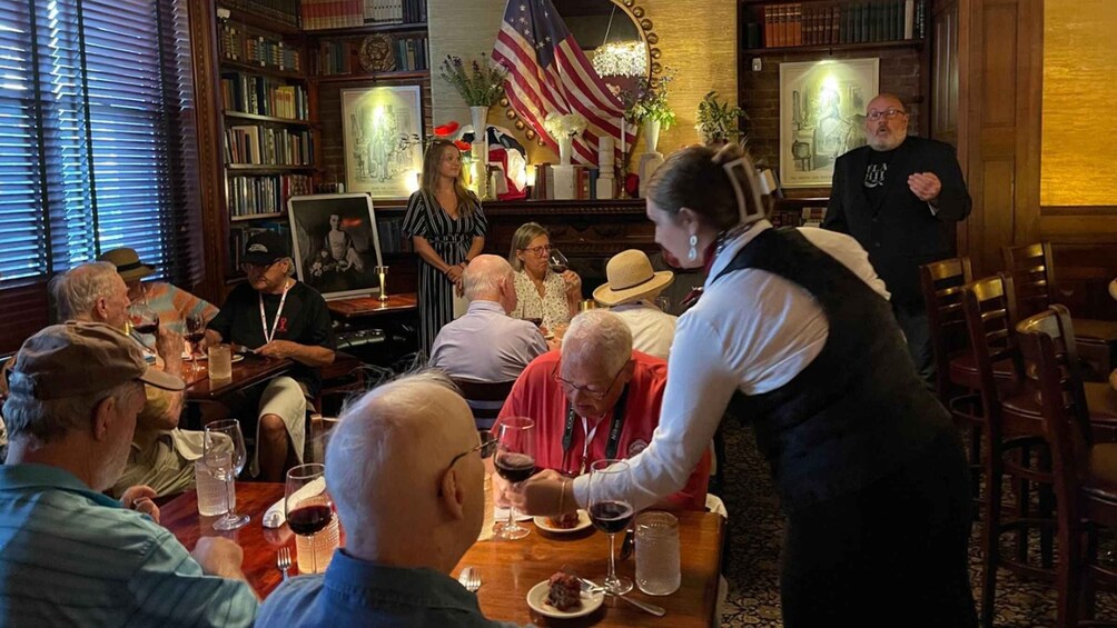 Eine Gruppe von Personen sitzt an einem Tisch in einem Restaurant, im Hintergrund weht eine amerikanische Flagge und steht ein Bücherregal. 3