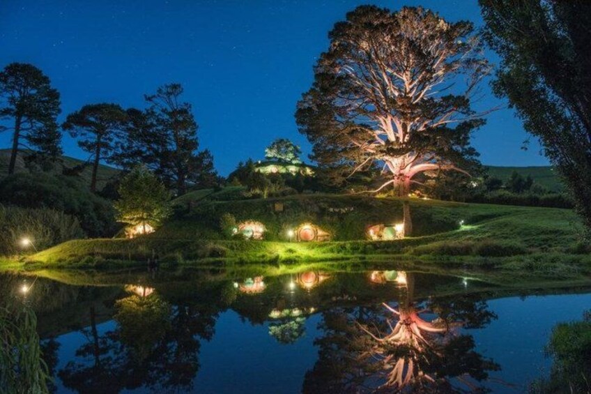 A nighttime landscape with a large illuminated tree, a pond reflecting the scene, and surrounding greenery. 3