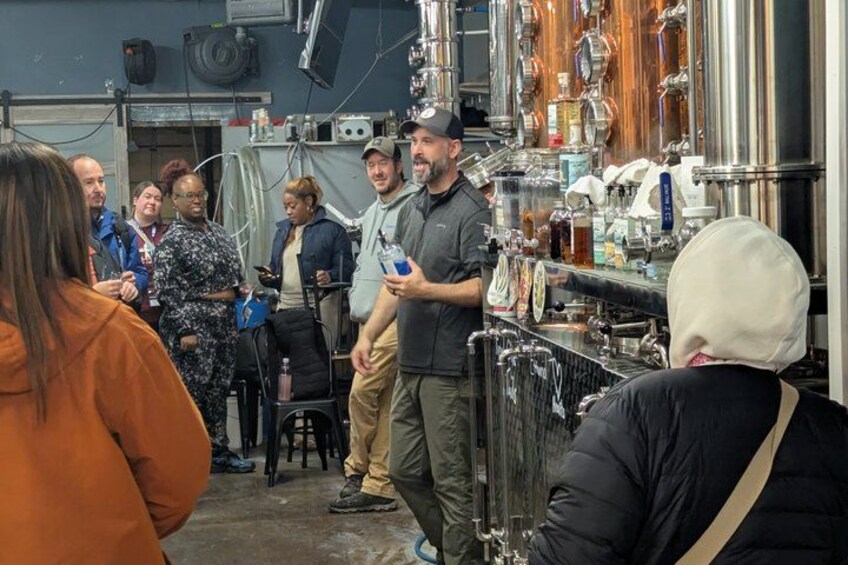 A group of people in a brewery setting with stainless steel tanks and brewing equipment. 1