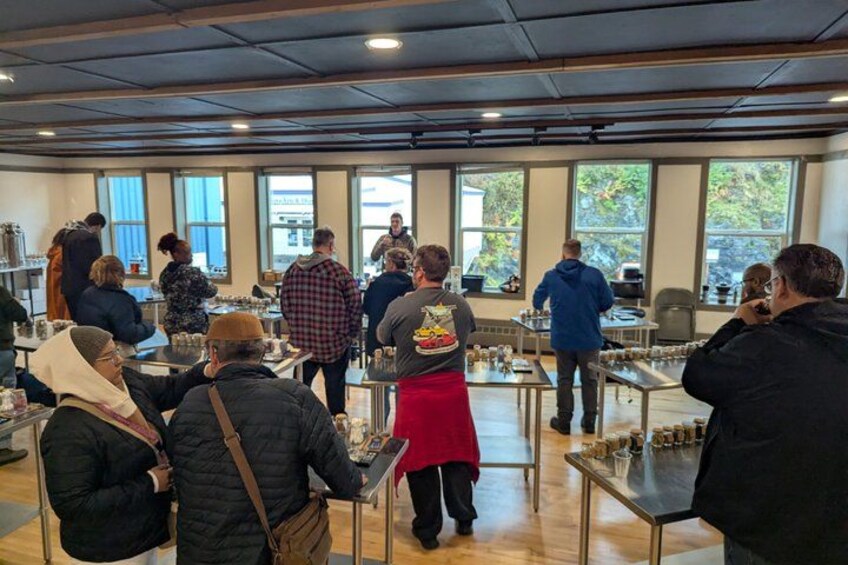 A group of people in a room with tables and chairs, some standing and some seated, engaged in conversation or activities. 3