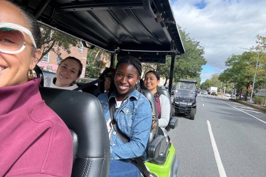 A group of people in a golf cart on a street with a vehicle in the background. 5