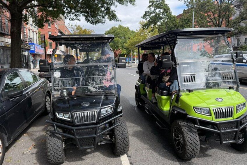 Two golf carts on a street with buildings and parked cars. 1