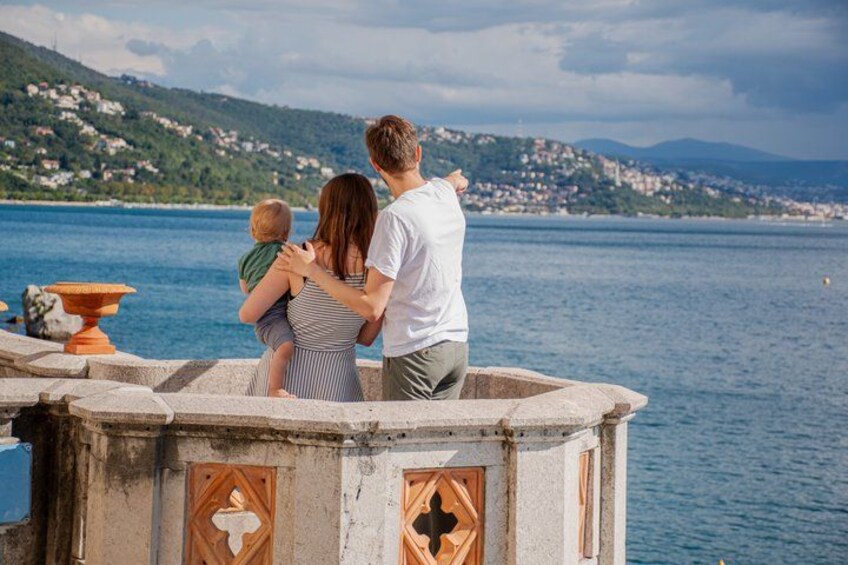 A family of three by a waterfront with a stone structure and a view of a distant town. 1