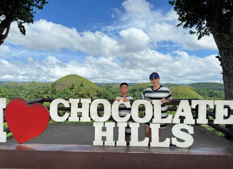 A sign with 'I ♥ CHOCOLATE HILLS' and a scenic view of green hills. 1